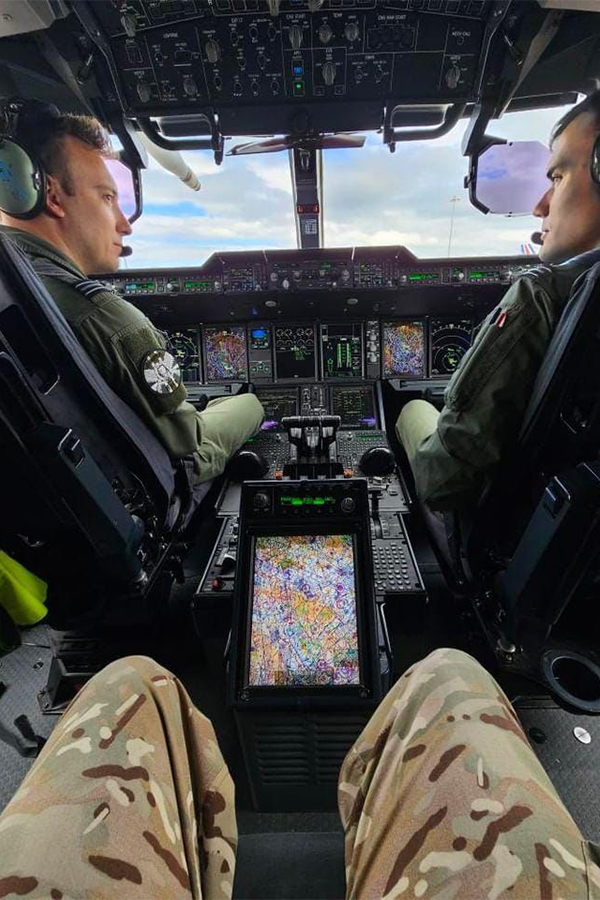 View from inside a military aircraft cockpit showing advanced flight controls and multiple digital displays, with two uniformed crew members seated at the controls and a person in camouflage trousers in the foreground. 
