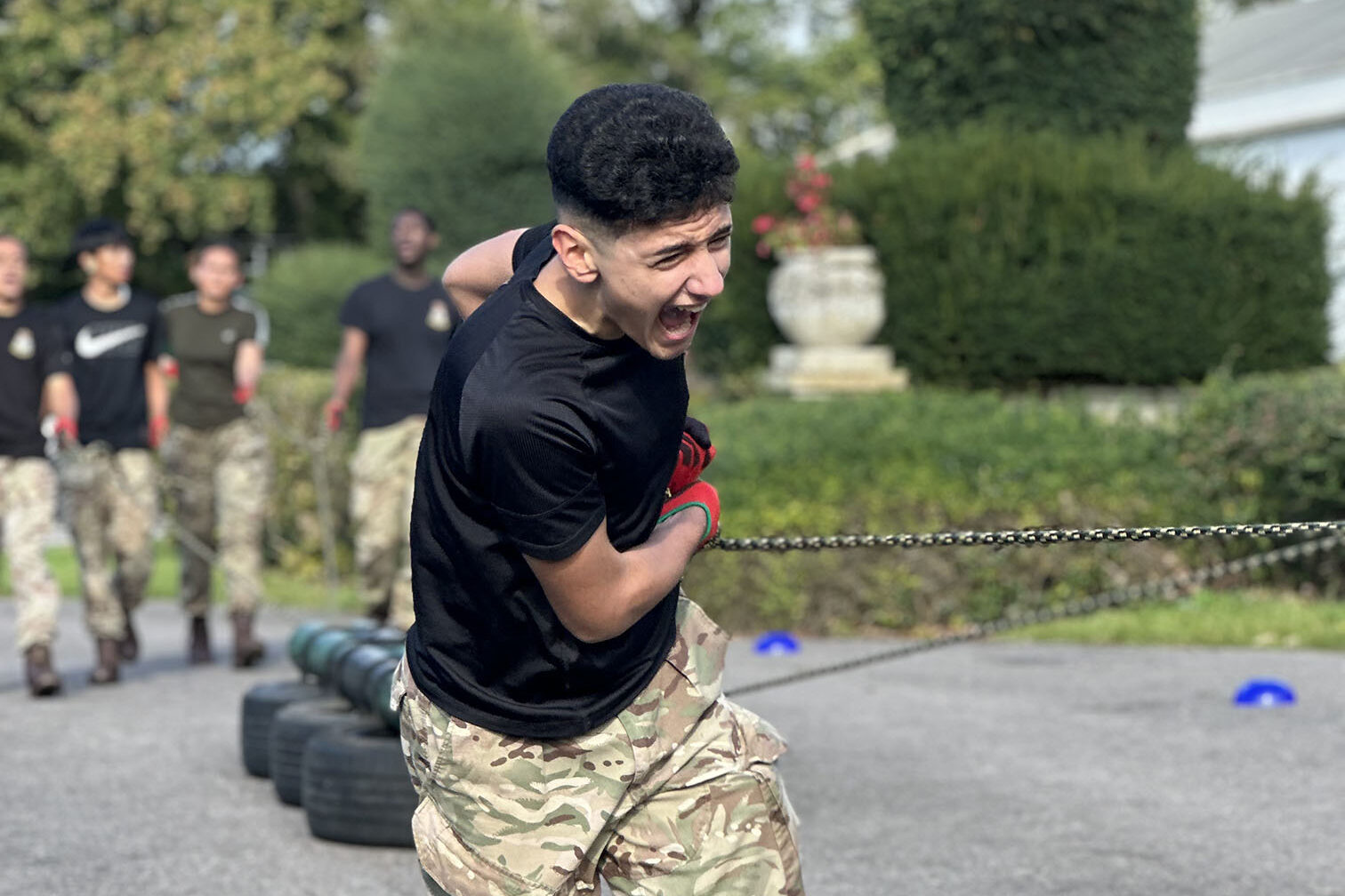 An Air Cadet in camouflage trousers and boots pulling a heavy chain attached to stacked tyres during an outdoor physical challenge, with other cadets in similar attire watching in the background. 
