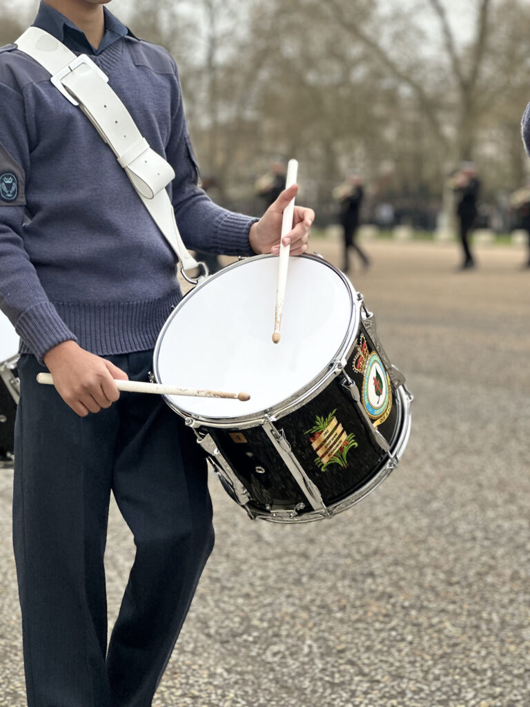 An Air Cadet wearing a blue uniform plays a large marching drum with two drumsticks during an outdoor parade or ceremonial event, with other participants visible in the background. 