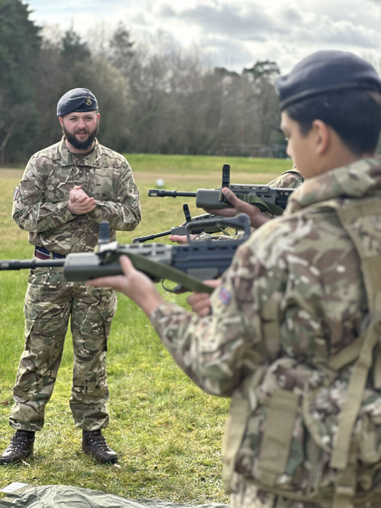 Air Cadets in camouflage uniforms holding training rifles during a field exercise, with an instructor standing in front giving guidance on a grassy outdoor range. 