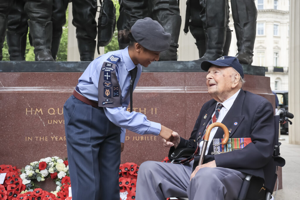 An Air Cadet in blue uniform shakes hands with a decorated veteran seated in a wheelchair at a memorial, with wreaths of red poppies laid at the base and an inscription honouring HM Queen Elizabeth II visible in the background. 