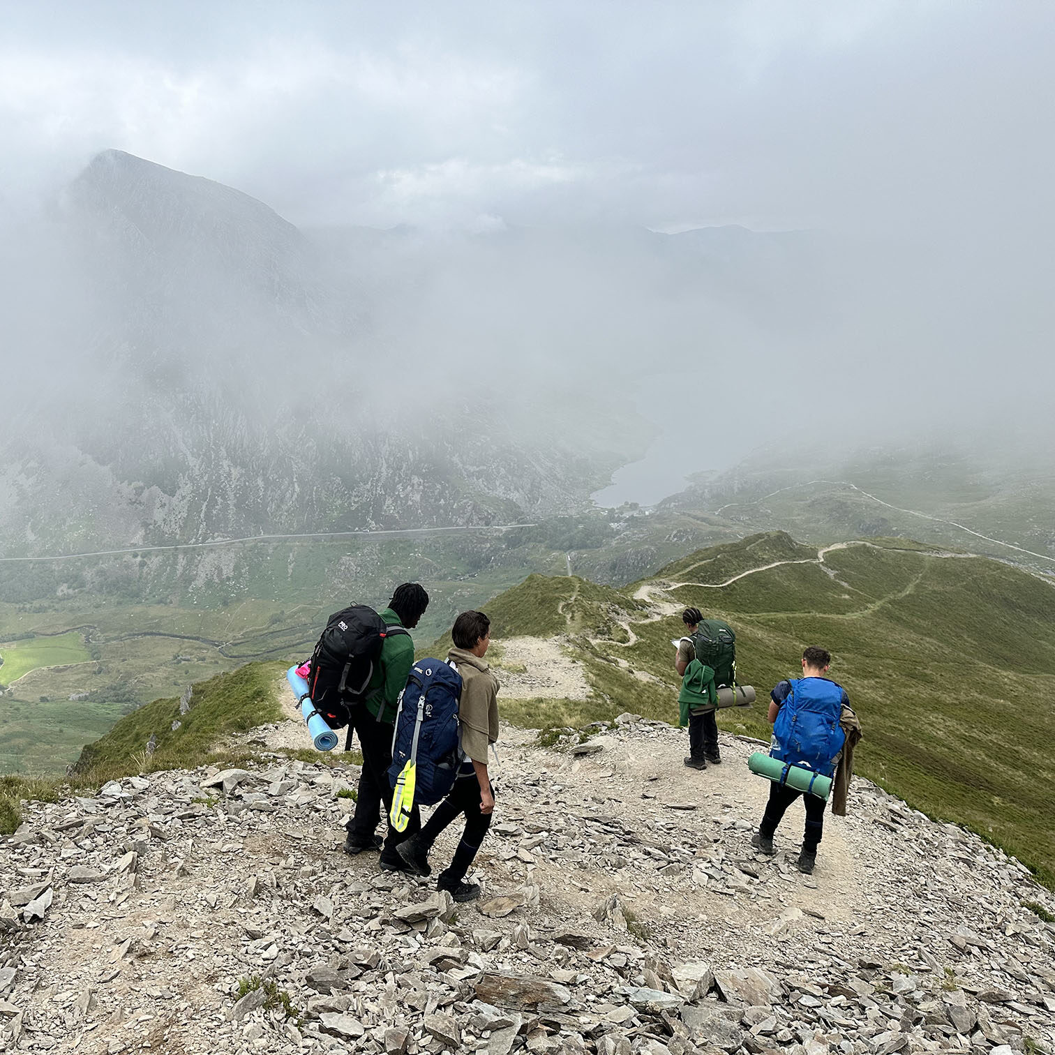 A group of Air Cadets hiking along a rocky mountain trail with large backpacks and camping gear, surrounded by misty peaks and a valley below, during an outdoor expedition. 