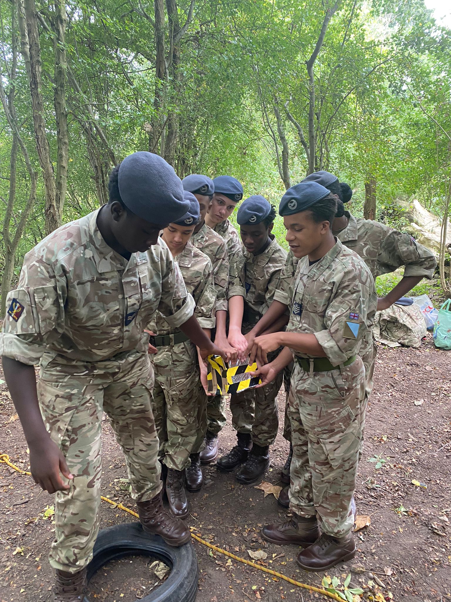 Air Cadets in camouflage uniforms and berets working together on a team-building exercise in a wooded area, holding a small object wrapped in black and yellow tape as part of an outdoor leadership activity. 
